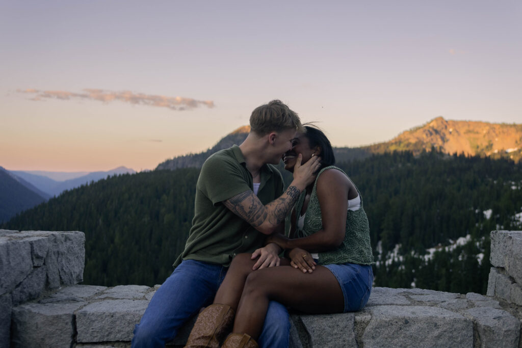 The couple celebrates being just married by exploring Mount Rainier together. the mountains are behind them as they share a kiss, sitting on a ledge.