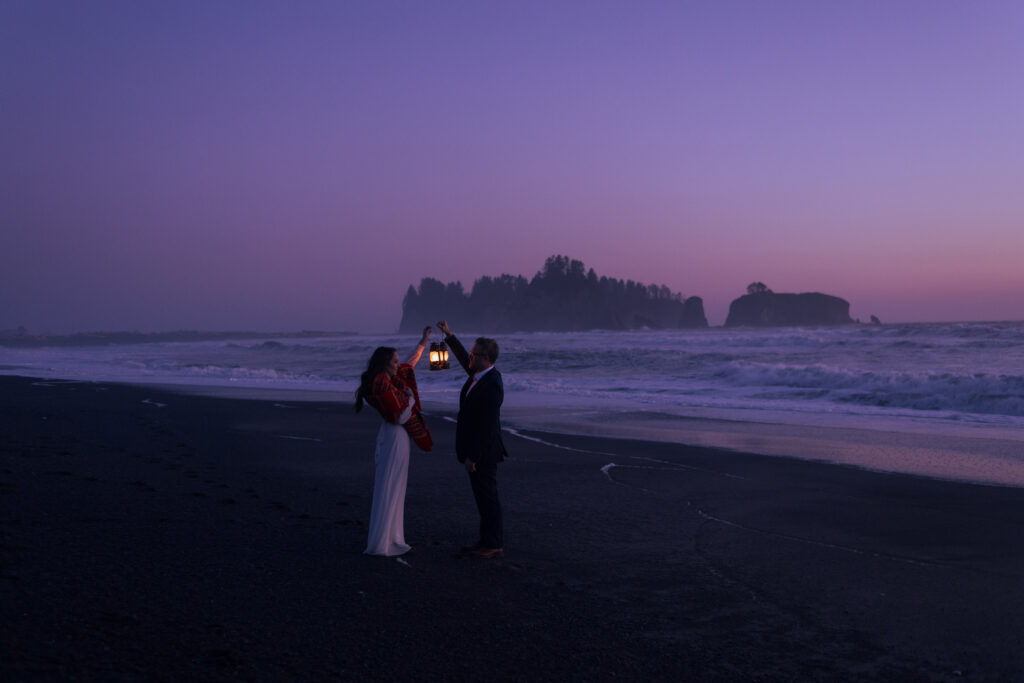 The sunset is bright pink while the bride and groom carry lanterns to light up their faces. The bride carries a blanket over her shoulders as the air becomes crispy cold.